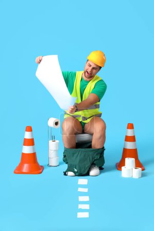 Téléchargez les photos : Travailleur assis sur un bol de toilette avec plan et cônes de circulation sur fond bleu - en image libre de droit