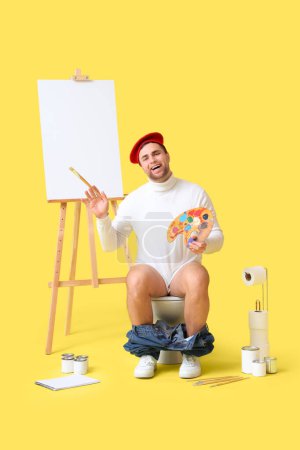 Téléchargez les photos : Artiste masculin avec palette de peinture et pinceau assis sur un bol de toilette sur fond jaune - en image libre de droit