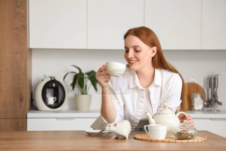 Beautiful redhead woman with cute white rat drinking tea in kitchen
