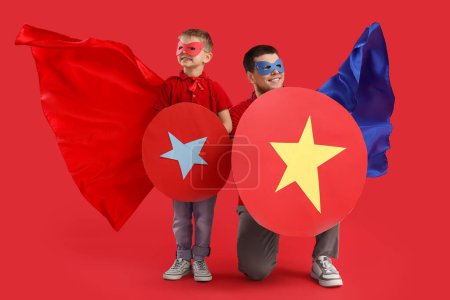 Photo for Little boy and his father in superhero costumes with paper shields on red background - Royalty Free Image