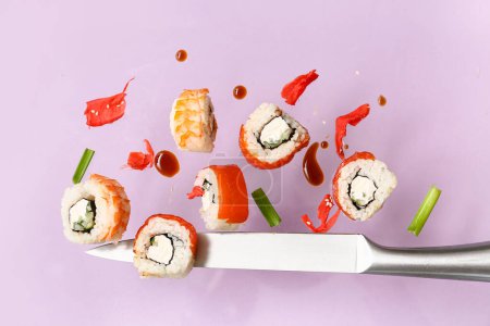 Deliciosos rollos de sushi voladores, jengibre, salsa y cuchillo sobre fondo lila