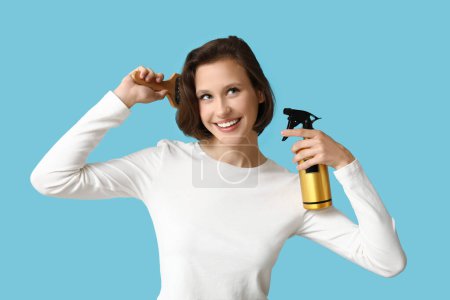 Beautiful young happy woman with bob hairstyle holding bottle of spray and brush on blue background