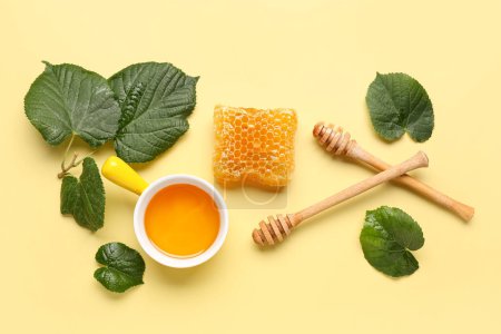 Bowl with tasty honey, honeycomb, wooden dippers and green leaves on beige background