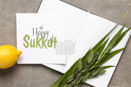 Four species (lulav, hadas, arava, etrog), open book and greeting card with text HAPPY SUKKOT on grey grunge background