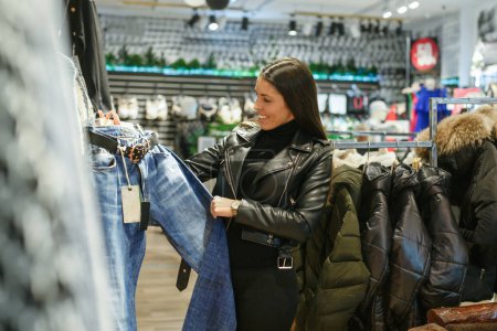 Téléchargez les photos : Adulte fille souriante acheter des pantalons dans le magasin de vêtements - en image libre de droit