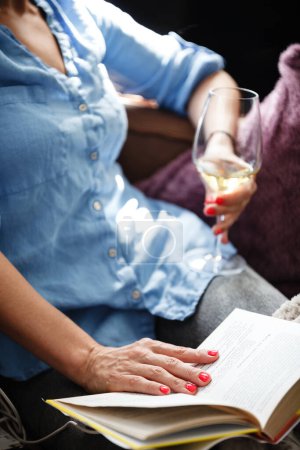 Téléchargez les photos : Concept de temps libre. Joyeux belle femme lit un livre et boit du vin assis sur un canapé à l'intérieur. Femme passer sa journée libre et se détendre à la maison seul - en image libre de droit