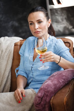 Téléchargez les photos : Concept de temps libre. Joyeux belle femme boit du vin blanc à partir d'un verre assis sur un canapé à l'intérieur. Femme passer sa journée libre et se détendre à la maison seul - en image libre de droit