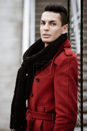 Photo for LGBTQ community lifestyle concept. Young homosexual man stands near glass. Handsome fashionable gay male model poses in cityscape outdoors. Wears red coat, gloves, and black scarf. - Royalty Free Image