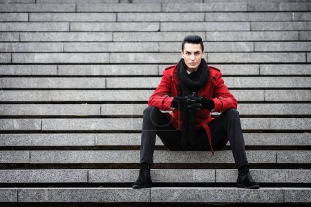 Photo for LGBTQ community lifestyle concept. Young homosexual man sits on a stairs. Handsome fashionable gay male model poses in cityscape outdoors. Wears red coat, gloves, and black scarf. - Royalty Free Image