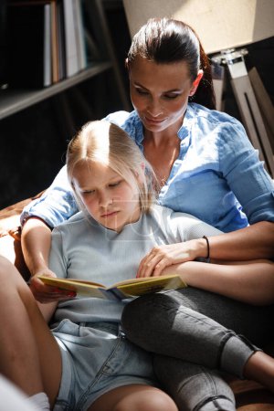 Téléchargez les photos : Concept de temps libre familial. Joyeux belle femme et adolescente lisant un livre assis sur un canapé à l'intérieur. Mère et fille passent une journée libre et apprennent à la maison - en image libre de droit
