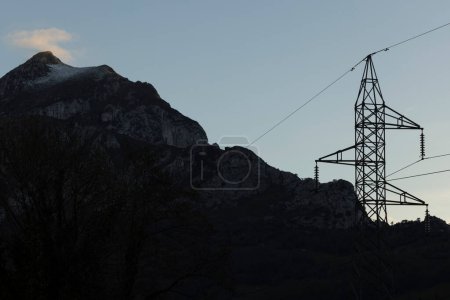 Electrical tower with wires next to high mountain peak with fog at sunset.