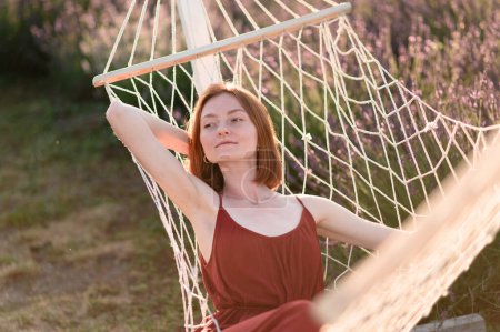 Photo for A red-haired young girl without makeup is resting on a hammock in a lavender field. Summer vacation and travel time. - Royalty Free Image