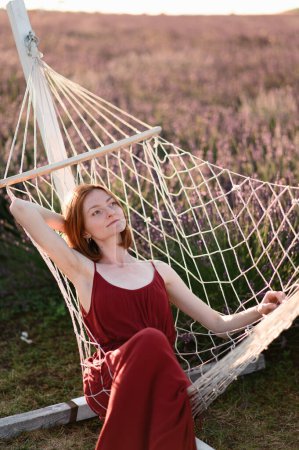 Photo for A red-haired young girl without makeup is resting on a hammock in a lavender field. Summer vacation and travel time. - Royalty Free Image