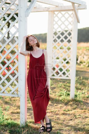 Photo for A red-haired young girl without makeup is resting near the gazebo in a flower field. Summer vacation and travel time. - Royalty Free Image