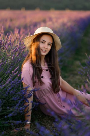 Photo for A cheerful young girl in a straw hat and a pink dress sits and rests among lavender bushes. Sunset. - Royalty Free Image