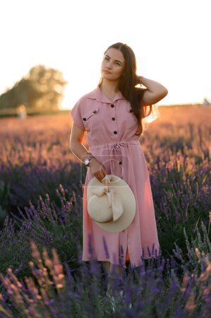 Photo for A cheerful young girl in a pink dress and a straw hat in her hands stands among lavender bushes. Sunset. - Royalty Free Image