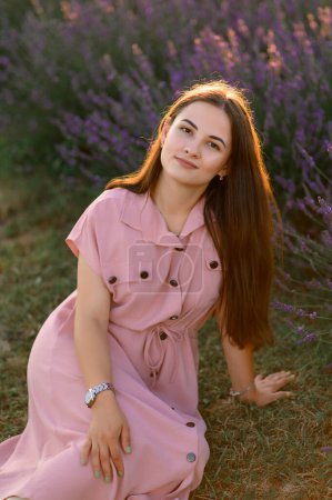 Photo for A cheerful young girl in a pink dress sits and rests among lavender bushes. Sunset. - Royalty Free Image
