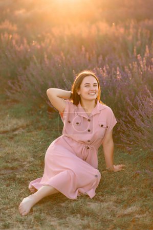 Photo for A cheerful young girl in a pink dress sits and rests among lavender bushes. Sunset. - Royalty Free Image