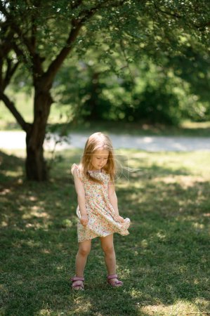 Photo for Little girl 3 years old in a summer sundress in the park. Summer time.The baby is playing with her dress. - Royalty Free Image