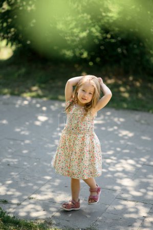 Photo for Little girl 3 years old in a summer sundress in the park. Summer time. The little girl raised her hands to her head. - Royalty Free Image