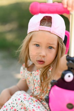 Photo for A little girl 3 years old in a pink cap rides a scooter. Summer time. Close up. - Royalty Free Image