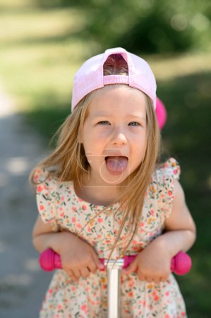 Photo for A little girl 3 years old in a pink cap rides a scooter. Summer time. The girl shows her tongue. - Royalty Free Image
