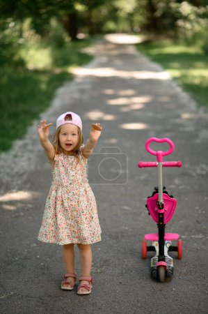 Photo for A little girl 3 years old in a pink cap rides a scooter. Summer time. The girl shows cookies. - Royalty Free Image
