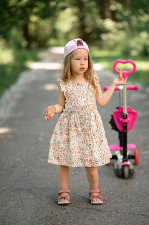 Photo for A little girl 3 years old in a pink cap rides a scooter. Summer time. - Royalty Free Image