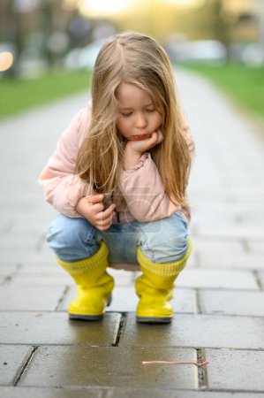 Photo for A little girl 3 years old walks in yellow rubber boots, after the rain and watches the earthworm. - Royalty Free Image