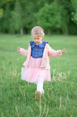 Photo for A little girl 3 years old walks in the park in a dress and a pink sweater. Summer time. The baby walks on the grass. - Royalty Free Image