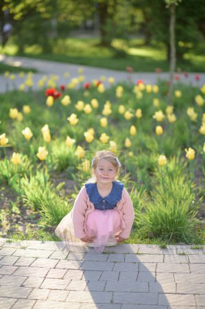 Photo for A little girl 3 years old walks in the park in a dress and a pink sweater. Summer time. The baby sits on a background of tulips. - Royalty Free Image