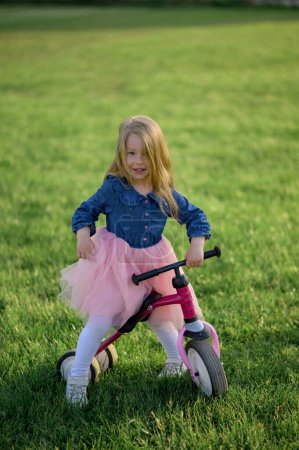 Photo for A happy beautiful little girl runs fast and rides her first bicycle without pedals on the grass on a warm summer day. Cute baby 3 years old. Side view. Learning to keep balance. - Royalty Free Image