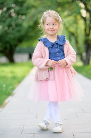 Photo for A little girl 3 years old walks in the park in a dress and a pink sweater. Summer time. The baby looks away with a smile. The girl crossed her legs. - Royalty Free Image