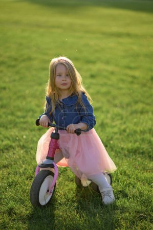 Photo for A happy beautiful little girl runs fast and rides her first bicycle without pedals on the grass on a warm summer day. Cute baby 3 years old. Side view. Learning to keep balance. - Royalty Free Image