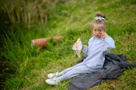 Photo for A little girl 3 years old sits in a suit on the grass in the park and eats pizza. The little girl is tired and rubs her eyes. - Royalty Free Image