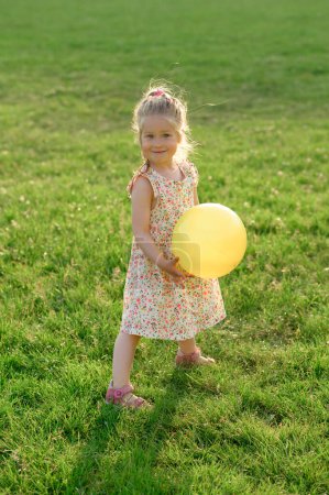 Photo for Little girl 3 years old in a summer dress in a clearing with an yellow ball in her hands. The girl is happy. - Royalty Free Image