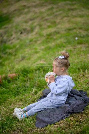 Photo for A little girl 3 years old sits in a suit on the grass in the park and eats pizza. The little girl is tired and rubs her eyes. - Royalty Free Image