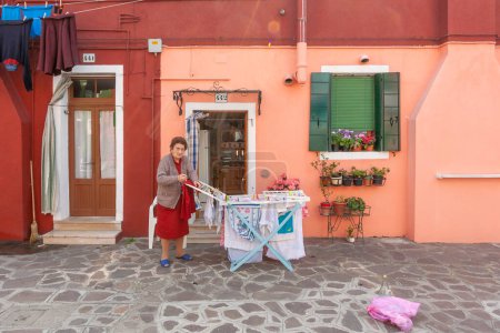 Téléchargez les photos : Burano, Italie - 10 avril 2007 : Une femme âgée met son linge sur un séchoir devant sa maison colorée à Burano, Venise, Italie. - en image libre de droit