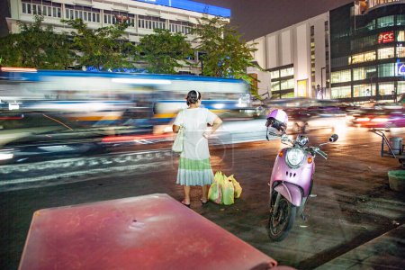 Téléchargez les photos : Bangkok, Thaïlande - 6 mai 2009 : femme après avoir fait du shopping attendre dans la rue Ratchadamri Road la nuit pour le prochain bus ou taxi avec le mouvement flou des voitures de passage. - en image libre de droit