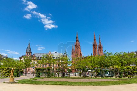 Foto de Wiesbaden, Alemania - 18 de mayo de 2023: vista desde la plaza del mercado hasta el antiguo ayuntamiento de Wiesbaden y la iglesia del mercado con árboles verdes en primer plano. - Imagen libre de derechos