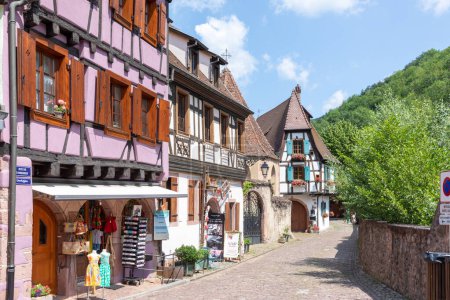 Foto de Kaysersberg, Francia - 23 de junio de 2023: Una pintoresca calle colorida de edificios de media madera con tiendas y cafeterías en el pueblo de Kaysersberg en la región de Vosges. - Imagen libre de derechos