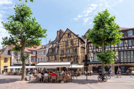 Foto de Colmar, Francia - 21 de junio de 2023: Plaza Dominicana en Colmar, Francia con casas de madera y turistas, disfrutando sentados en los cafés en un día de verano. - Imagen libre de derechos