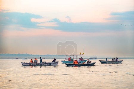 Téléchargez les photos : Varanasi, Inde - 24 février 2024 : de vieux bateaux en bois au Gange dans la lumière du matin avec des gens qui regardent le culte brûlant des hindous morts. - en image libre de droit