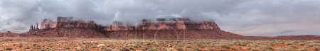 A panorama of the mountain range hosting the famous Three Sisters spires in Monument Valley, Arizona on the left of the image during a cloudy, rainy day.