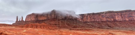 A panorama of the mountain range hosting the famous Three Sisters spires in Monument Valley, Arizona on the left of the image during a cloudy, rainy day.