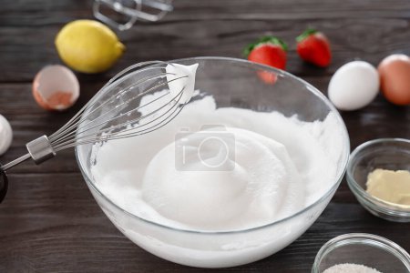 Close-up of a whisk and whipped egg whites to stiff peaks, a step in the preparation of breakfast dough or tender pancakes. White whipped foam of egg whites, bakery preparation. Wooden table