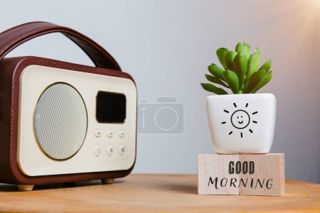 Un bonjour chaleureux sur des blocs de bois, un succulent charmant dans un pot de soleil souriant, et une radio nostalgique, créant un début de journée édifiant sur une table en bois.
