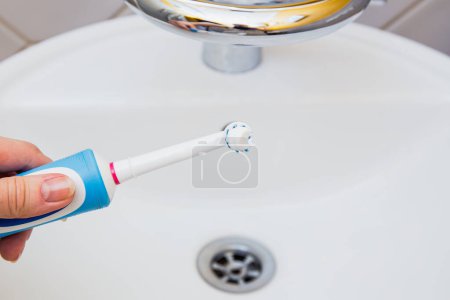 Téléchargez les photos : Brosse à dents électrique à main avec dentifrice dans la salle de bain près du robinet en céramique. routine quotidienne d'hygiène dentaire - en image libre de droit