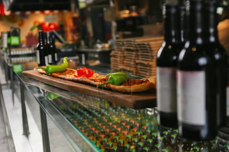 A rustic wooden board presenting pizza with peppers. Bottles of wine line the bar backdrop