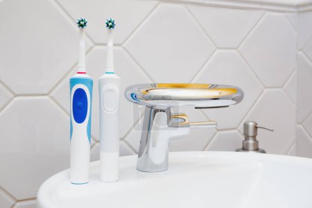 Two electric toothbrushes placed beside, family toothbrushes, modern sink with a rceramic faucet in a tiled bathroom.